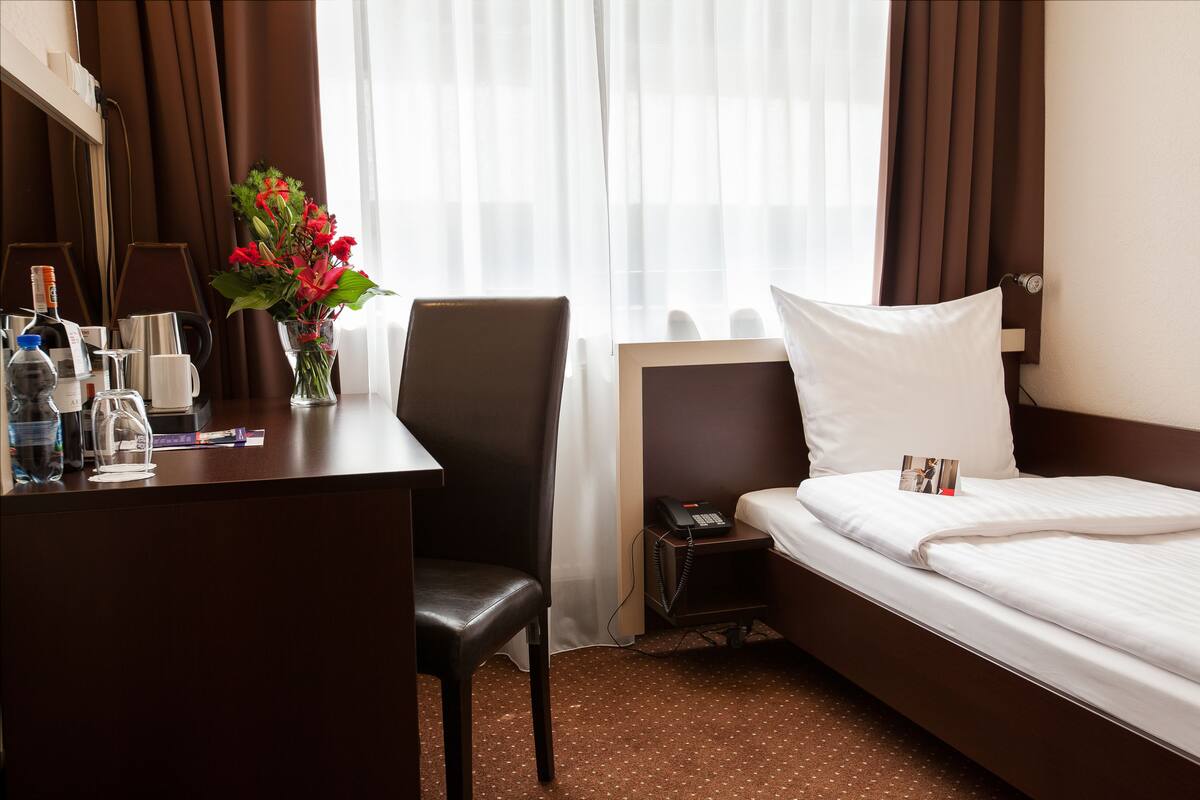 A hotel room with a bed, a desk with a lamp, a chair, a vase with flowers, and a telephone.