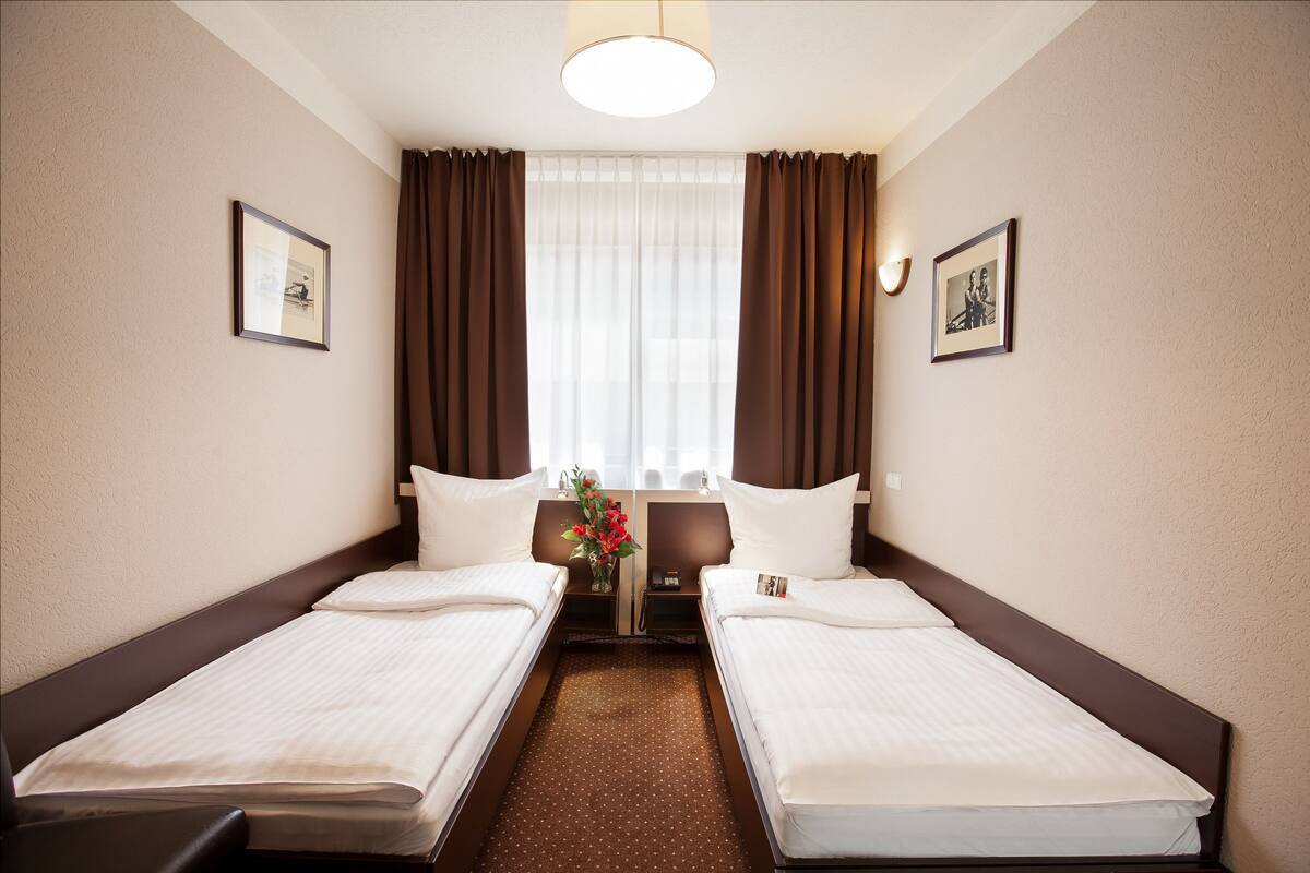 A hotel room with two single beds, a large window with curtains, a nightstand with a vase of flowers, and framed pictures on the walls.