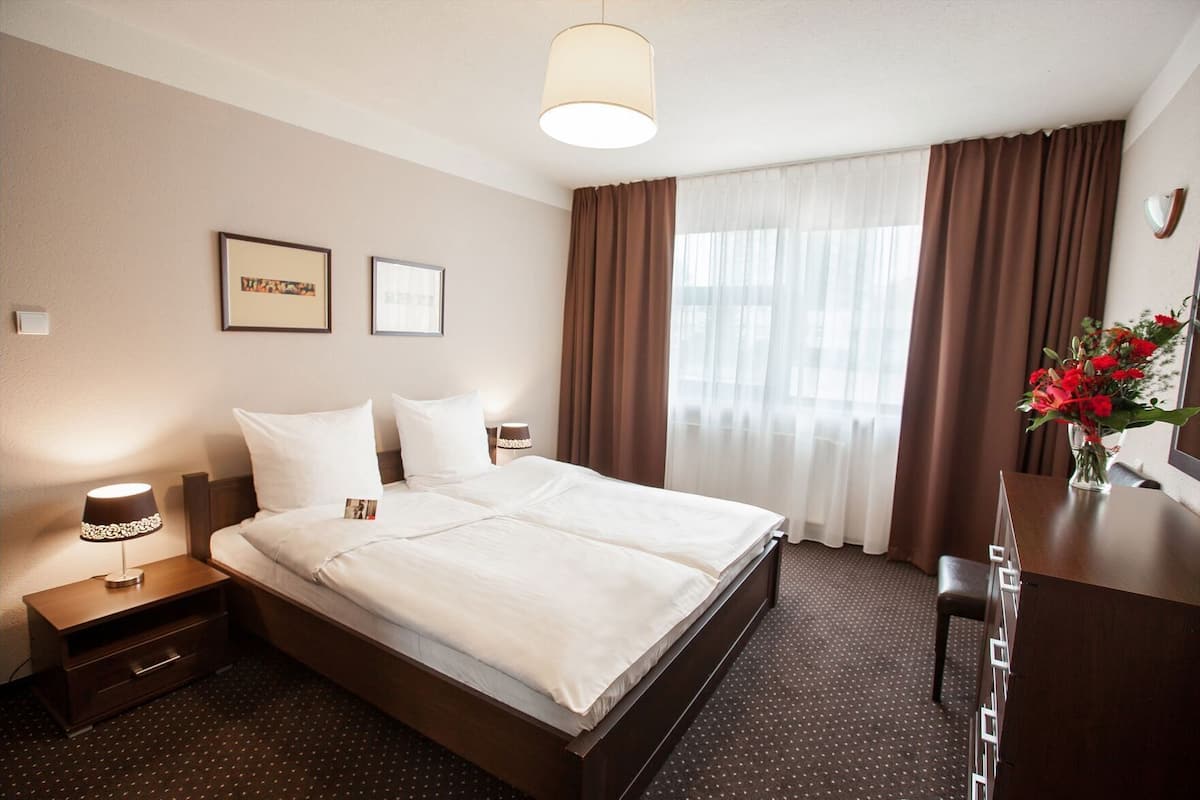 A hotel room with a bed, bedside tables, a desk, a lamp, curtains, and a window.
