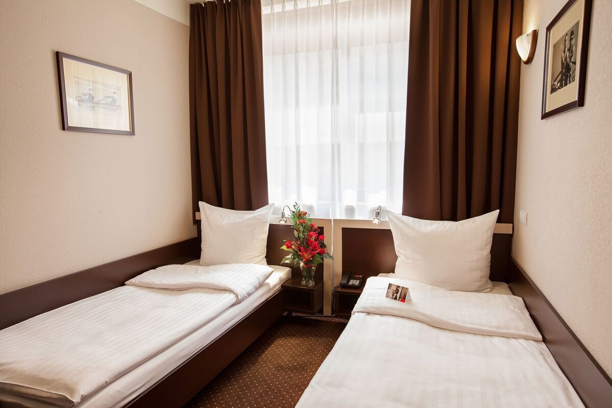 Una habitación de hotel con dos camas individuales, una mesita de noche con un teléfono, un jarrón con flores y un cuadro en la pared.