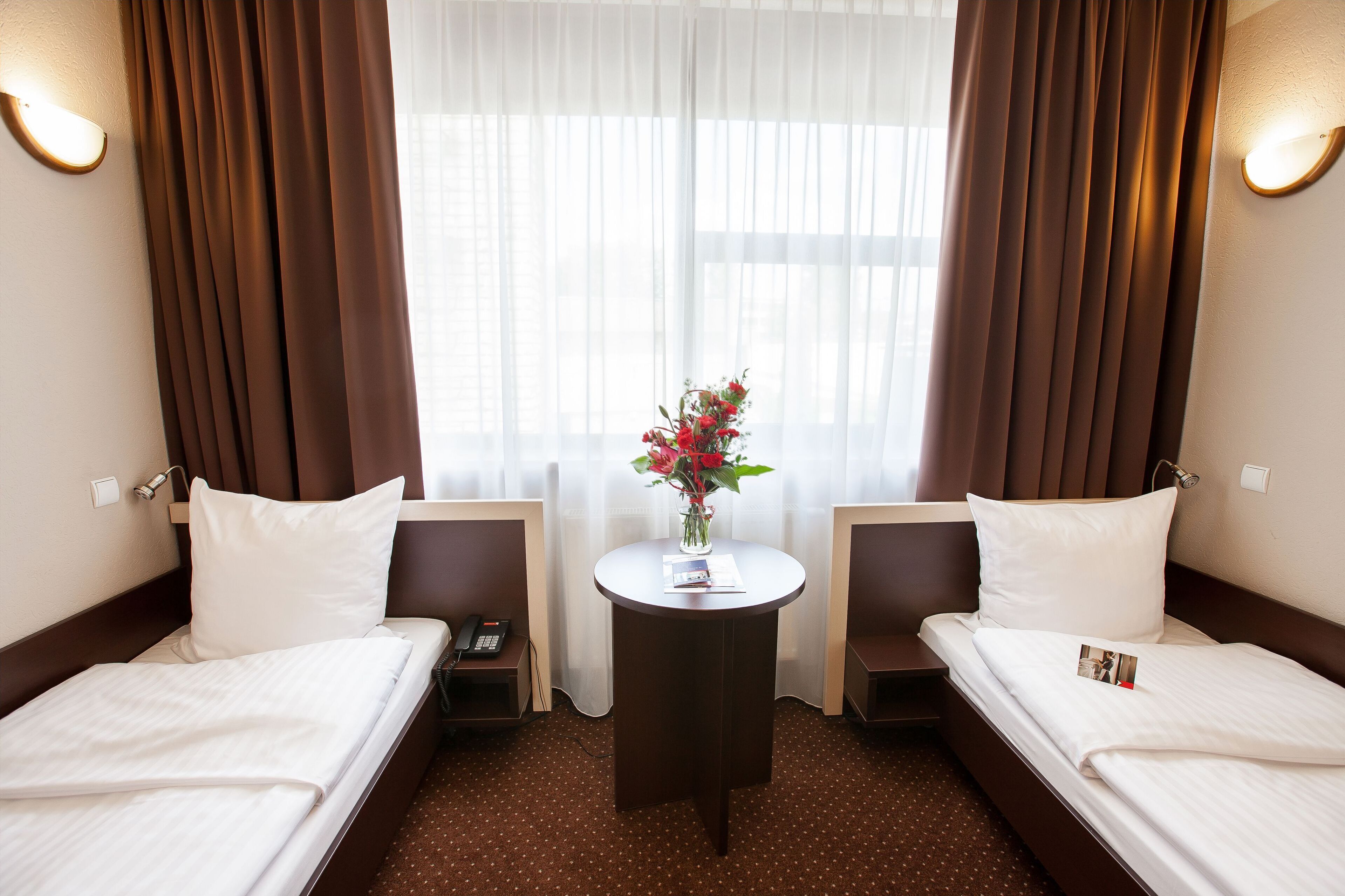 Habitación de hotel con dos camas, una mesita con un florero y una ventana con cortinas.