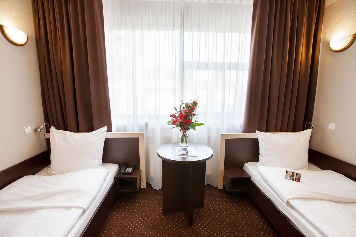 A hotel room with two beds, a small table with a vase of flowers, and a window with curtains.
