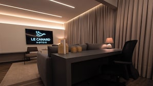 A modern hotel room with a flat-screen TV displaying the hotel name, a sofa, a desk with a lamp, and a chair. - Hotéis Le Canard (Lages)