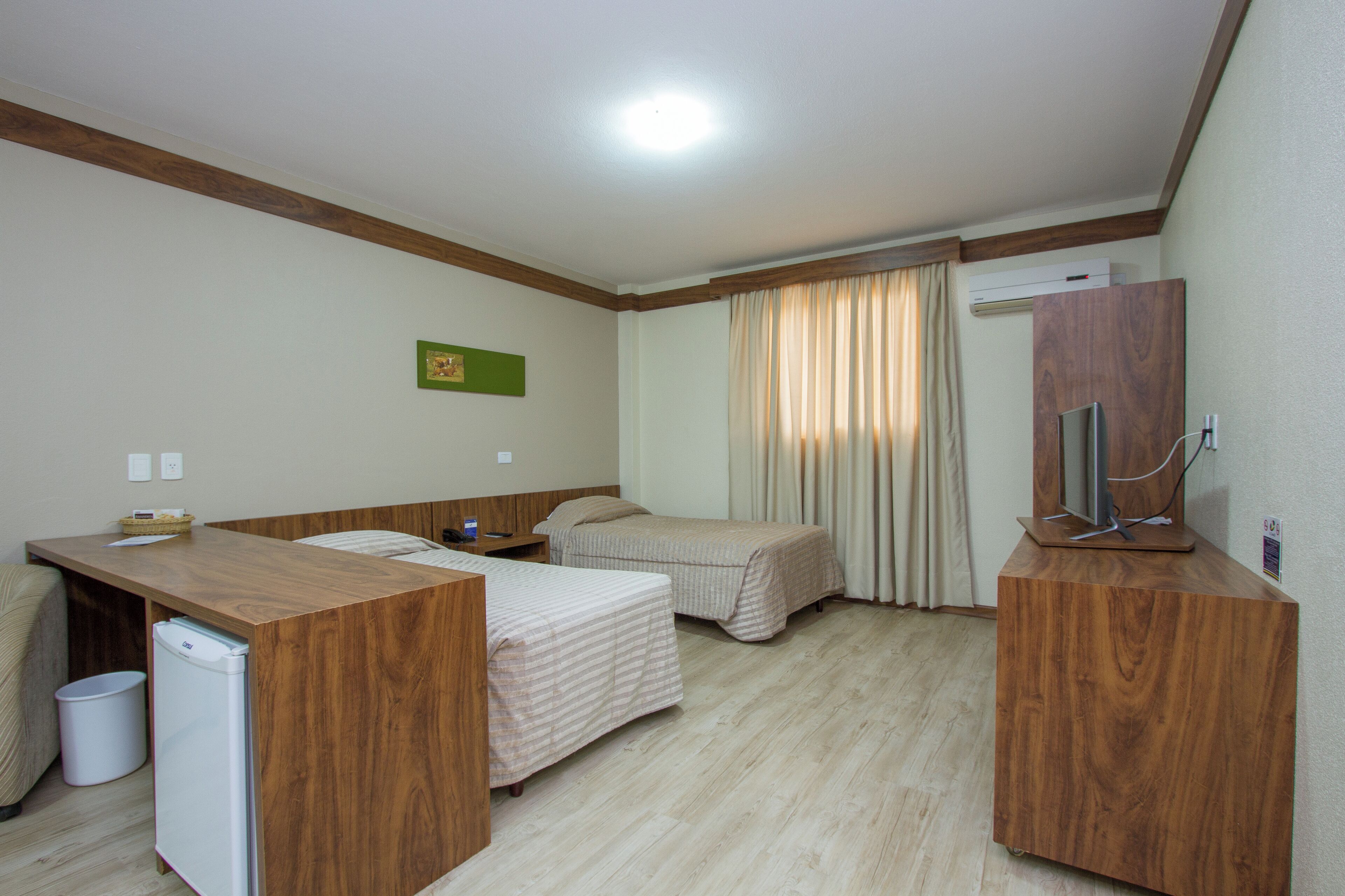 A hotel room with a wooden desk, two beds, a TV, and an air conditioning unit.
