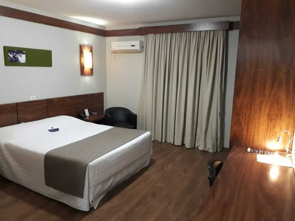 Quarto de hotel com cama, escrivaninha, cadeira, ar condicionado fixado na parede e janela com cortinas.