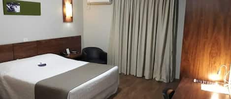 A hotel room with a bed, a desk, a chair, a wall-mounted air conditioner, and a window with curtains.