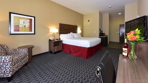 A hotel room with a bed, a desk, a chair, a lamp, a framed picture, a TV, and a vase with flowers.