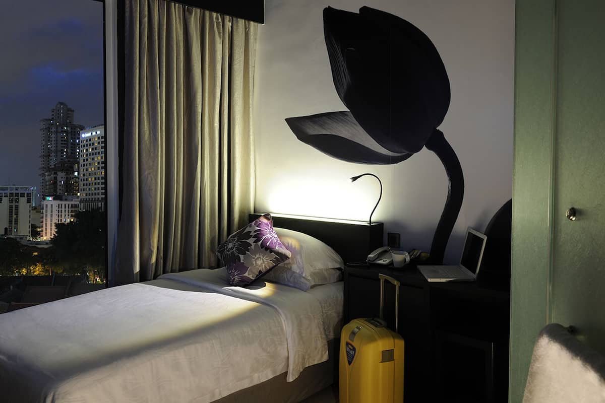 A hotel room with a bed, a nightstand with a lamp, a suitcase, and a view of the cityscape.