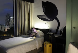A hotel room with a bed, a nightstand with a lamp, a suitcase, and a view of the cityscape.