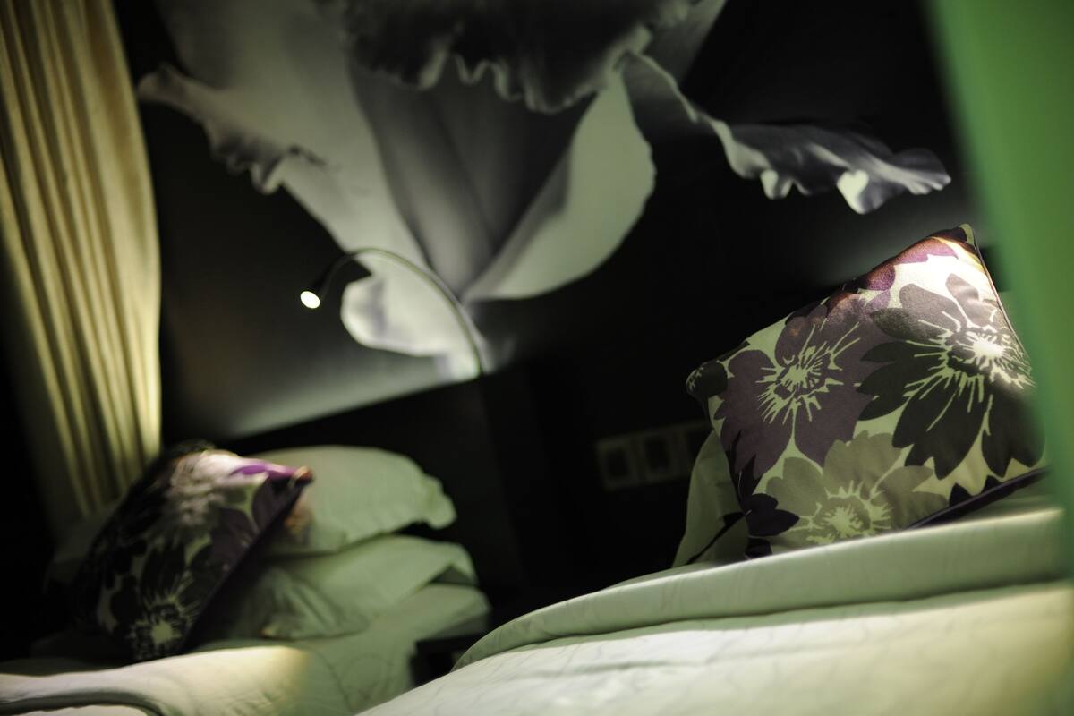 A bed with a floral pillow and a blanket, a wall with a large, dark artwork, and a partially visible curtain.