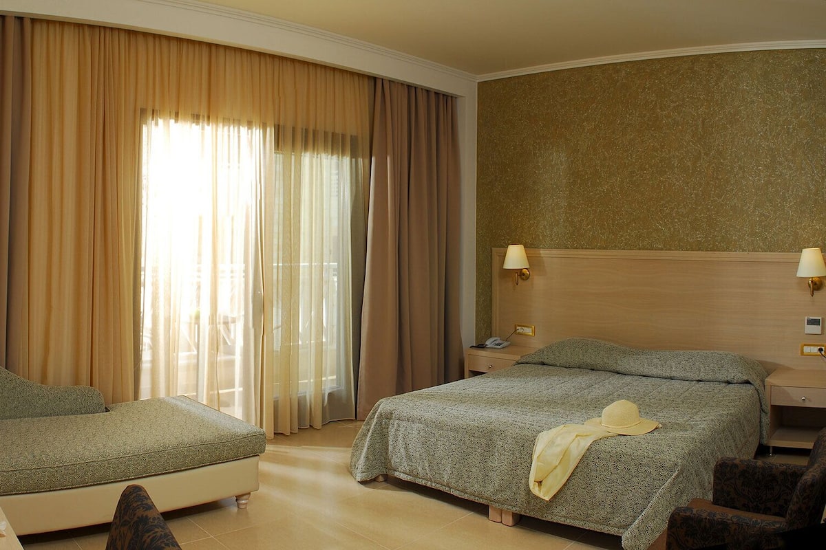 Une chambre d’hôtel avec un lit, une chaise, une table de chevet avec une lampe et une grande fenêtre avec des rideaux.