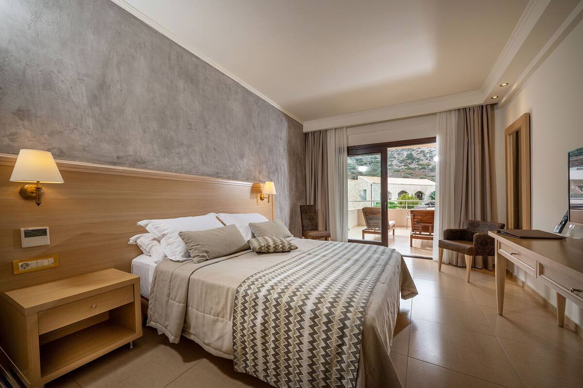 Une chambre d’hôtel comprenant un lit, des lampes de chevet, une table de nuit en bois, un bureau avec une chaise et une vue sur l’extérieur grâce à une porte vitrée.