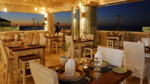 Un restaurante con varias mesas y sillas, grandes ventanales que ofrecen vista al océano, y un ambiente cálido con vigas de madera y luces colgantes.