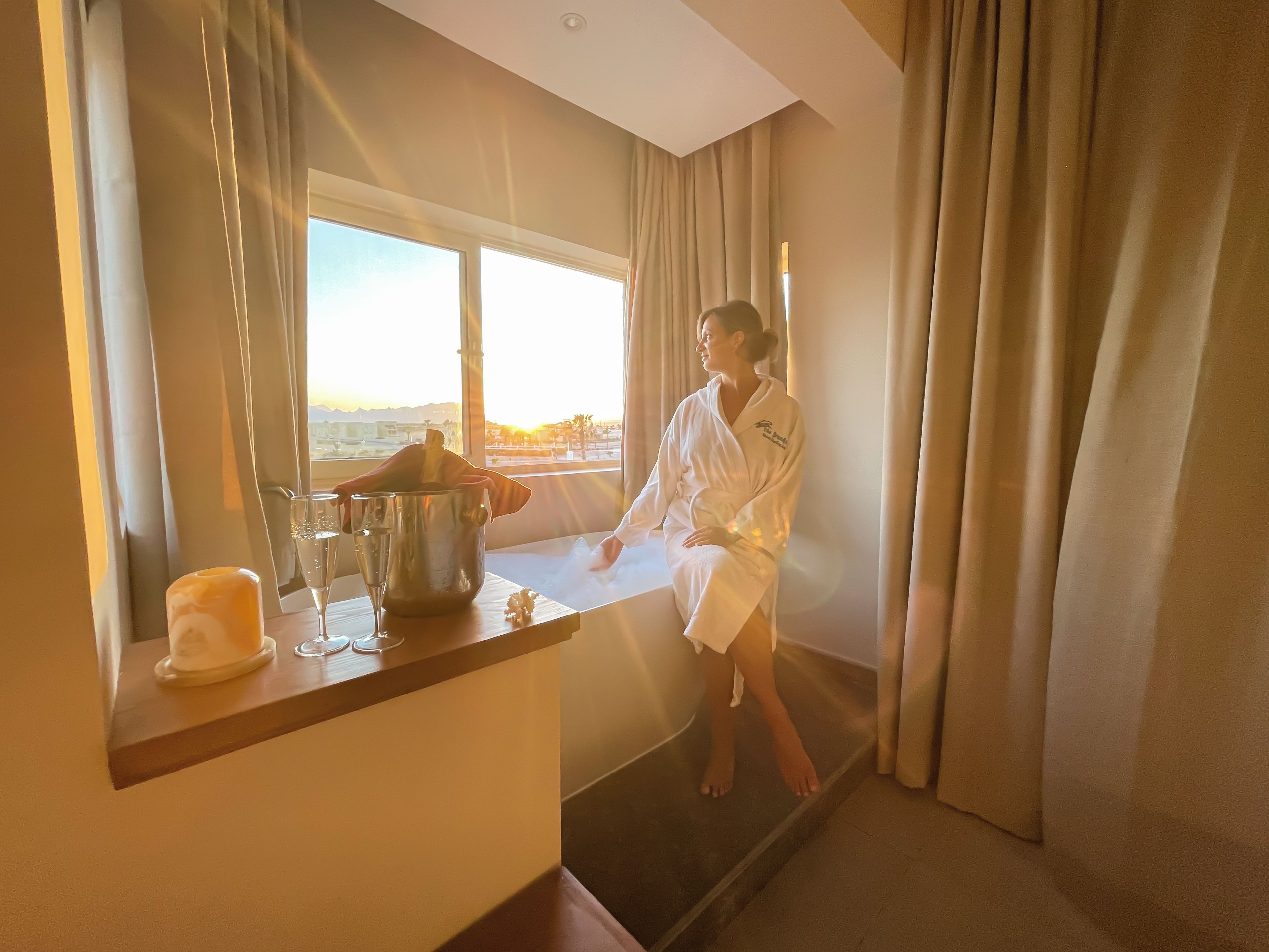 Una persona en un albornoz blanco sentada junto a una ventana con vista al atardecer.