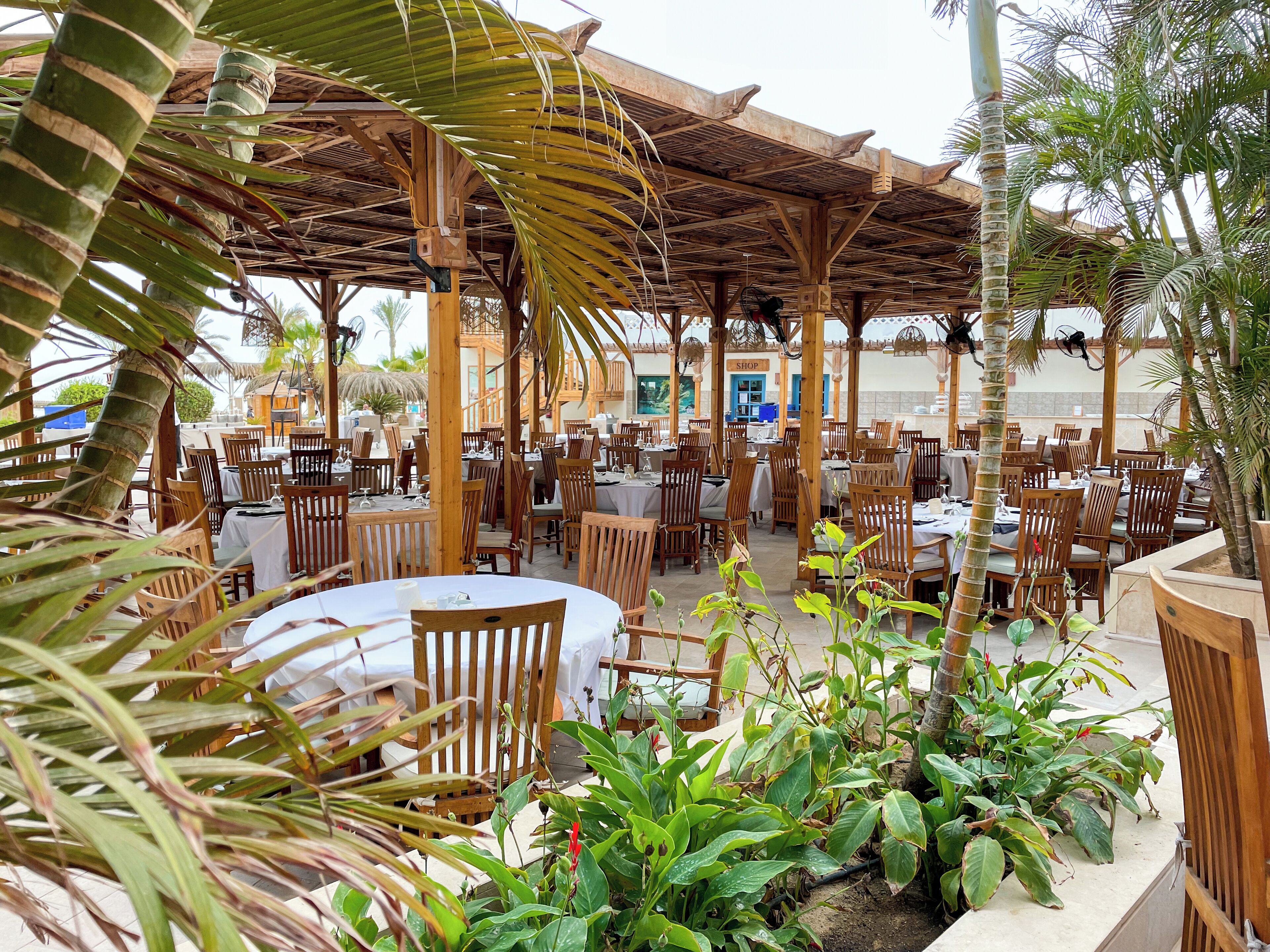 An outdoor restaurant with wooden tables and chairs, surrounded by tropical plants and a wooden pergola.