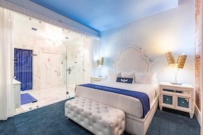 A bedroom with a large bed, a glass shower enclosure, a nightstand, and a wall-mounted light fixture.