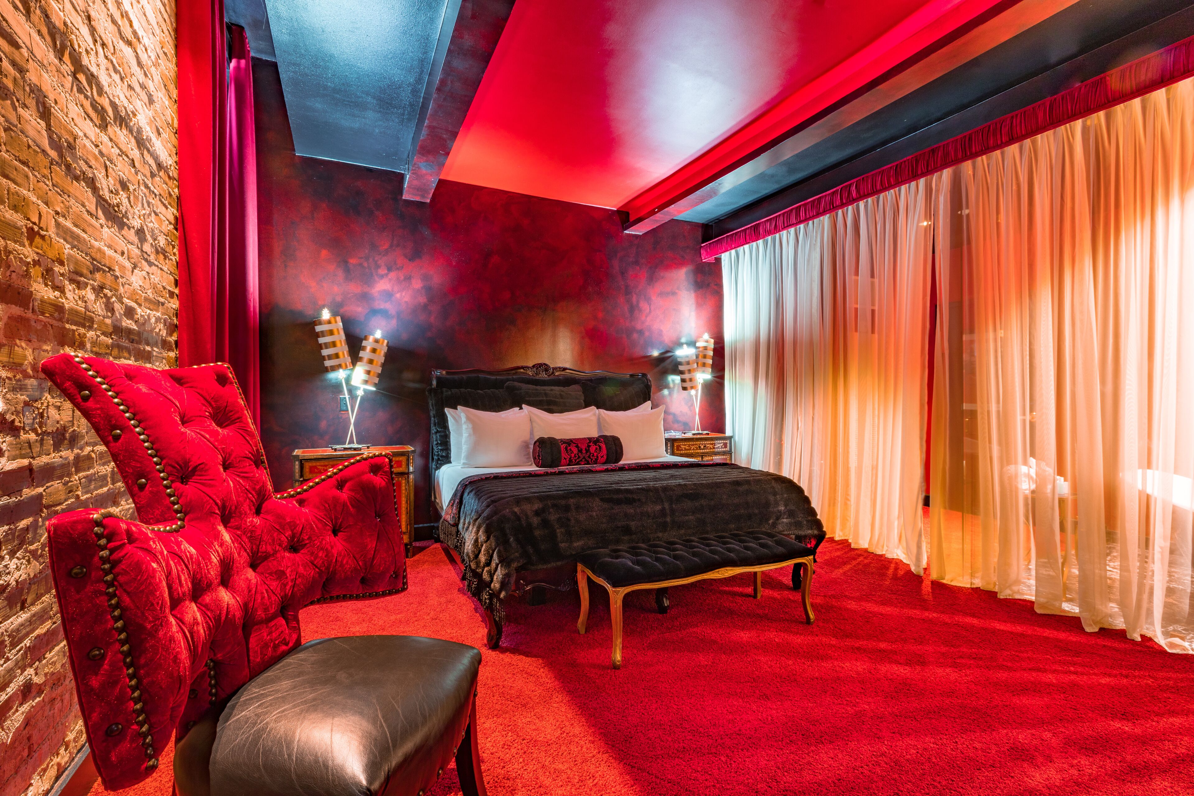 A room with a brick wall, a red upholstered chair, a bed with a black headboard, and a red carpet.