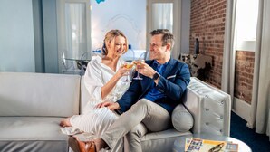 A couple sitting on a grey sofa in a room with a brick wall and a window. - The Saint Hotel, New Orleans, French Quarter (New Orleans)
