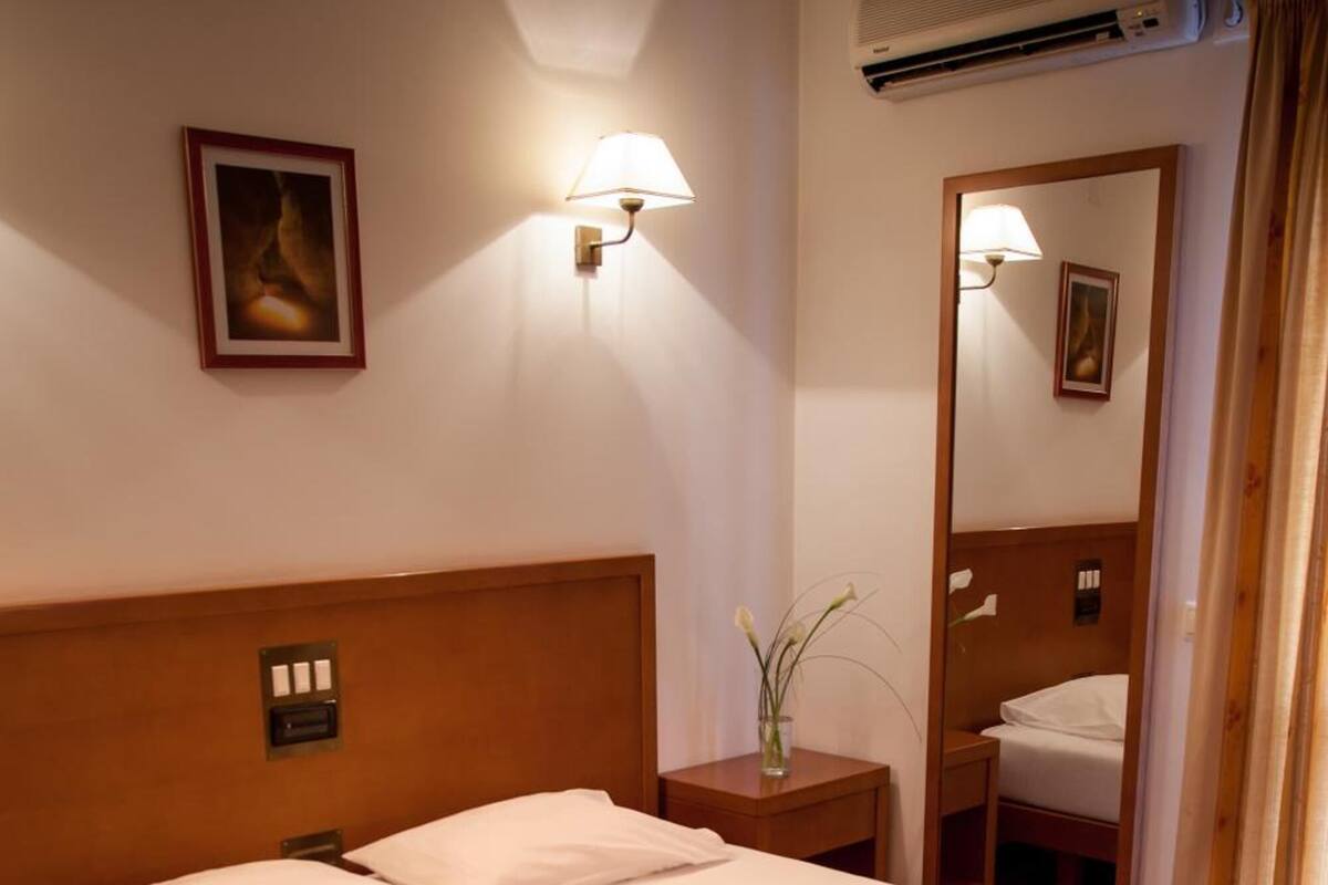 A hotel room with a bed, bedside table, lamp, mirror, and an air conditioning unit.