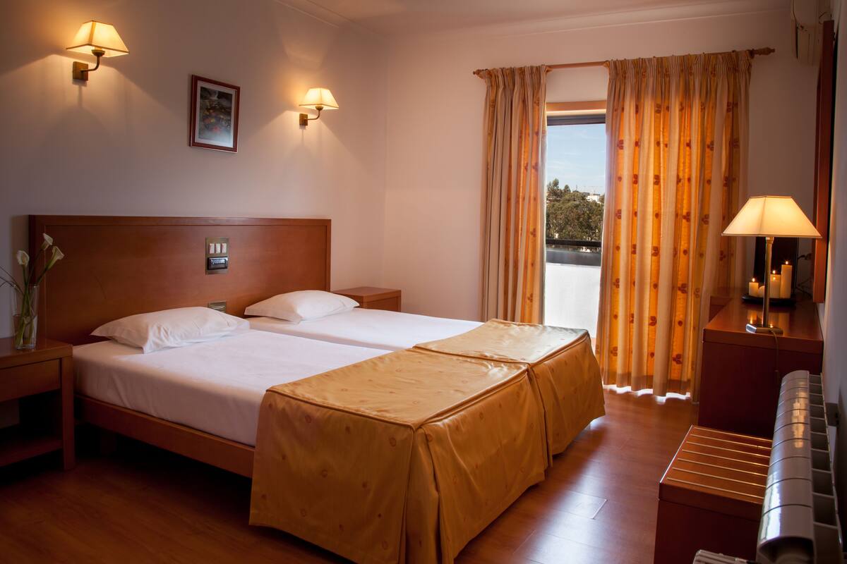 A hotel room with two beds, a piano, a desk with a lamp, and a view of the outdoors.