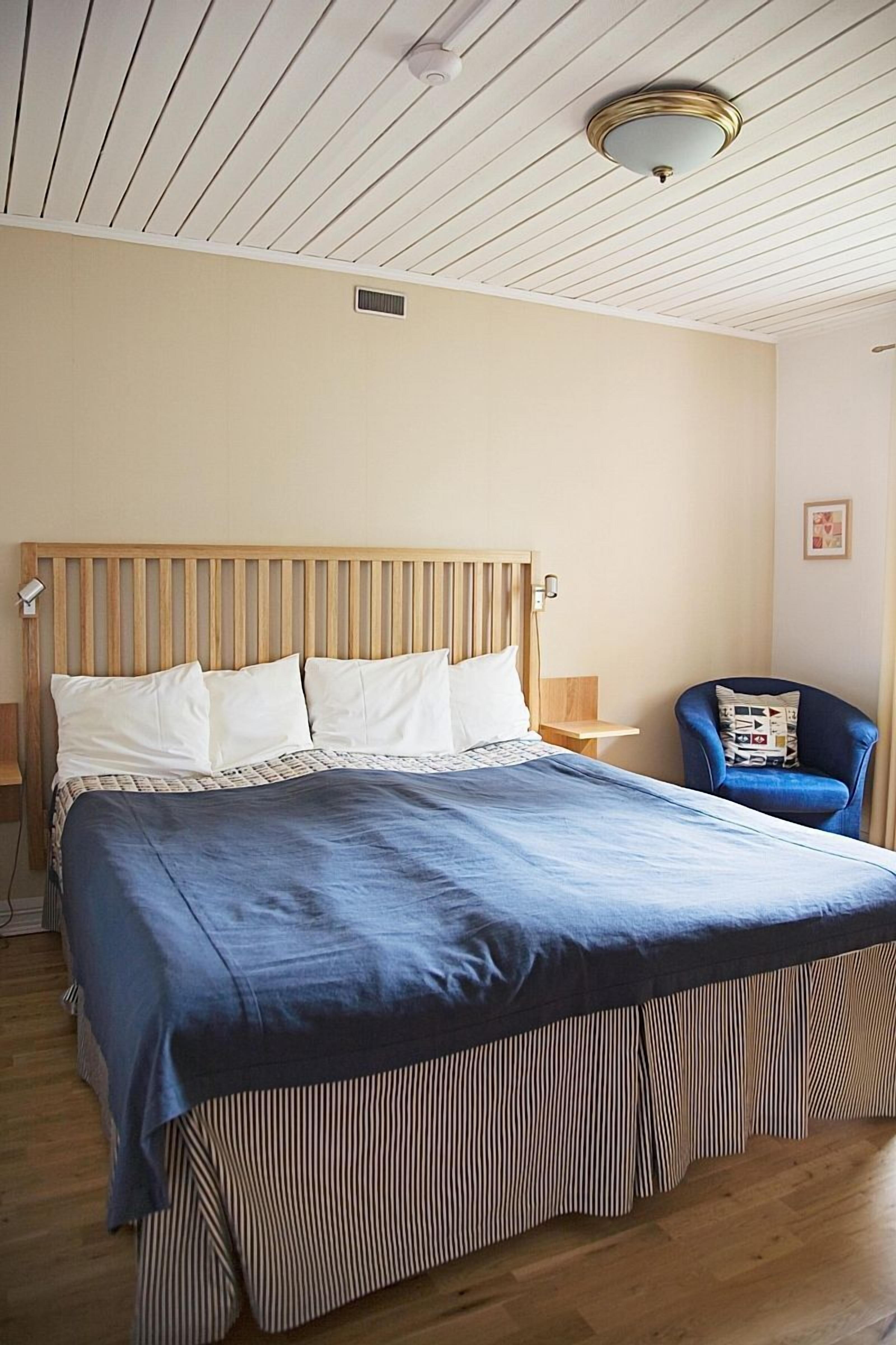 Un dormitorio con cabecera de madera, una cama grande con colcha azul, una silla con cojín estampado y una lámpara en el techo.