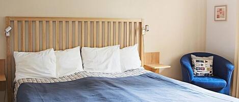 Un dormitorio con cabecera de madera, una cama grande con colcha azul, una silla con cojín estampado y una lámpara en el techo.
