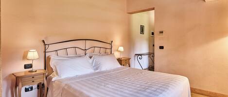 Una cama bien hecha con sábanas blancas, una mesita de noche con lámpara y una mesita auxiliar de madera.