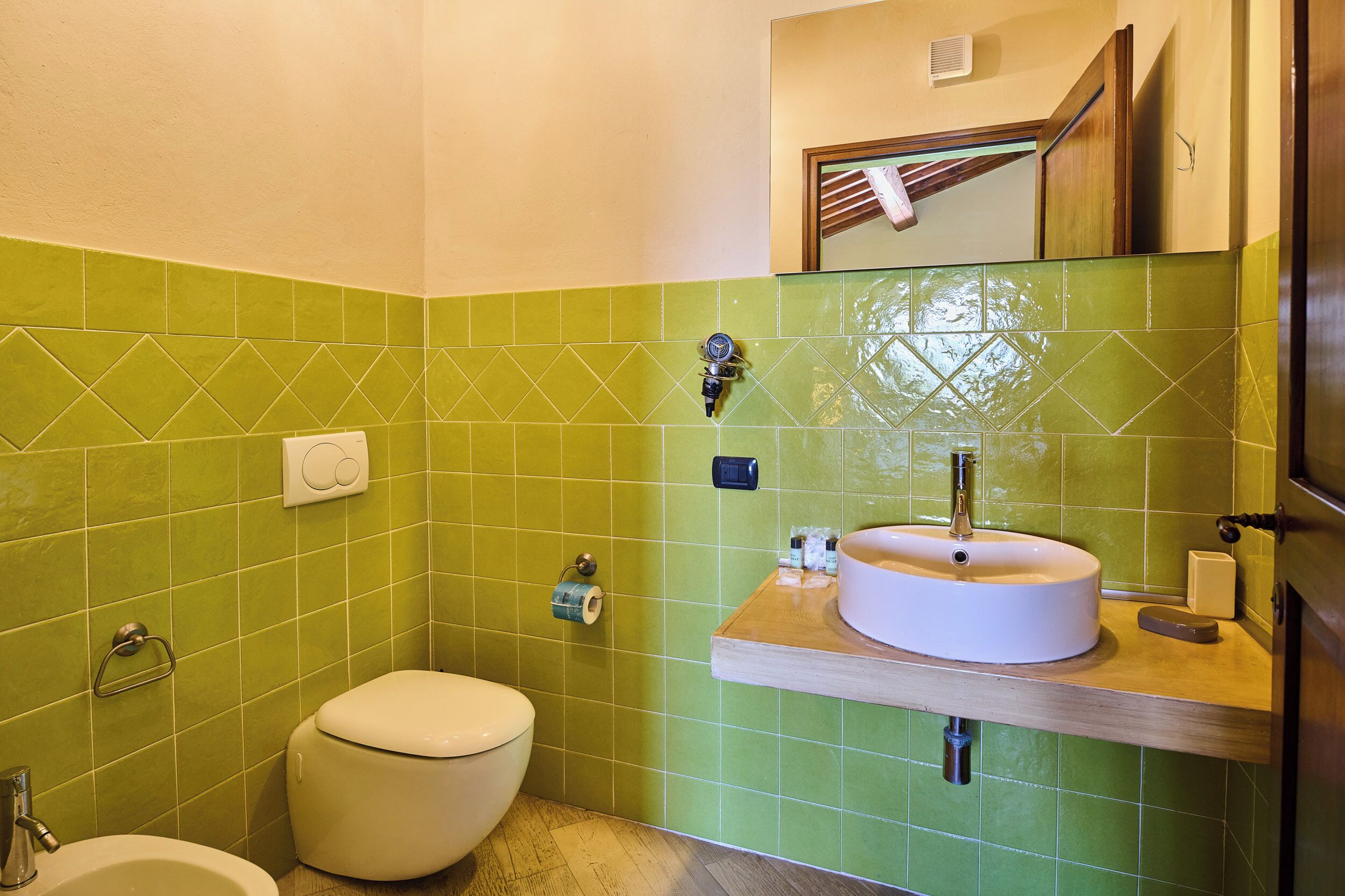 A bathroom with green tiles, a white toilet, a sink with a mirror above it, and a wooden countertop.
