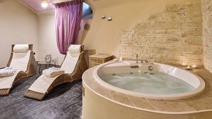 Een wellnessruimte met een groot bad, twee rieten lounge fauteuils, een tafeltje met kaarsen en een stenen muur.