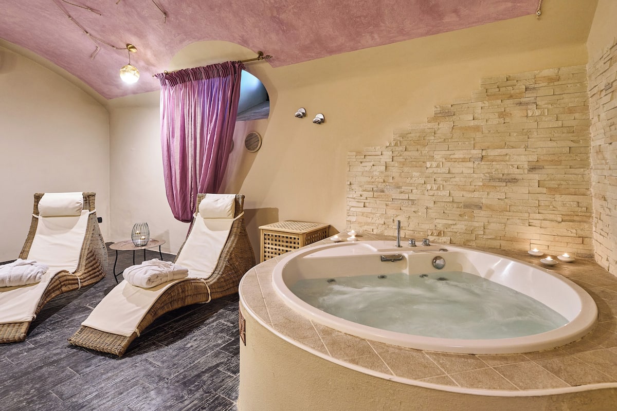 Une salle de spa dotée d’une grande baignoire, de deux fauteuils de relaxation en osier, d’une petite table avec des bougies et d’un mur en pierre.