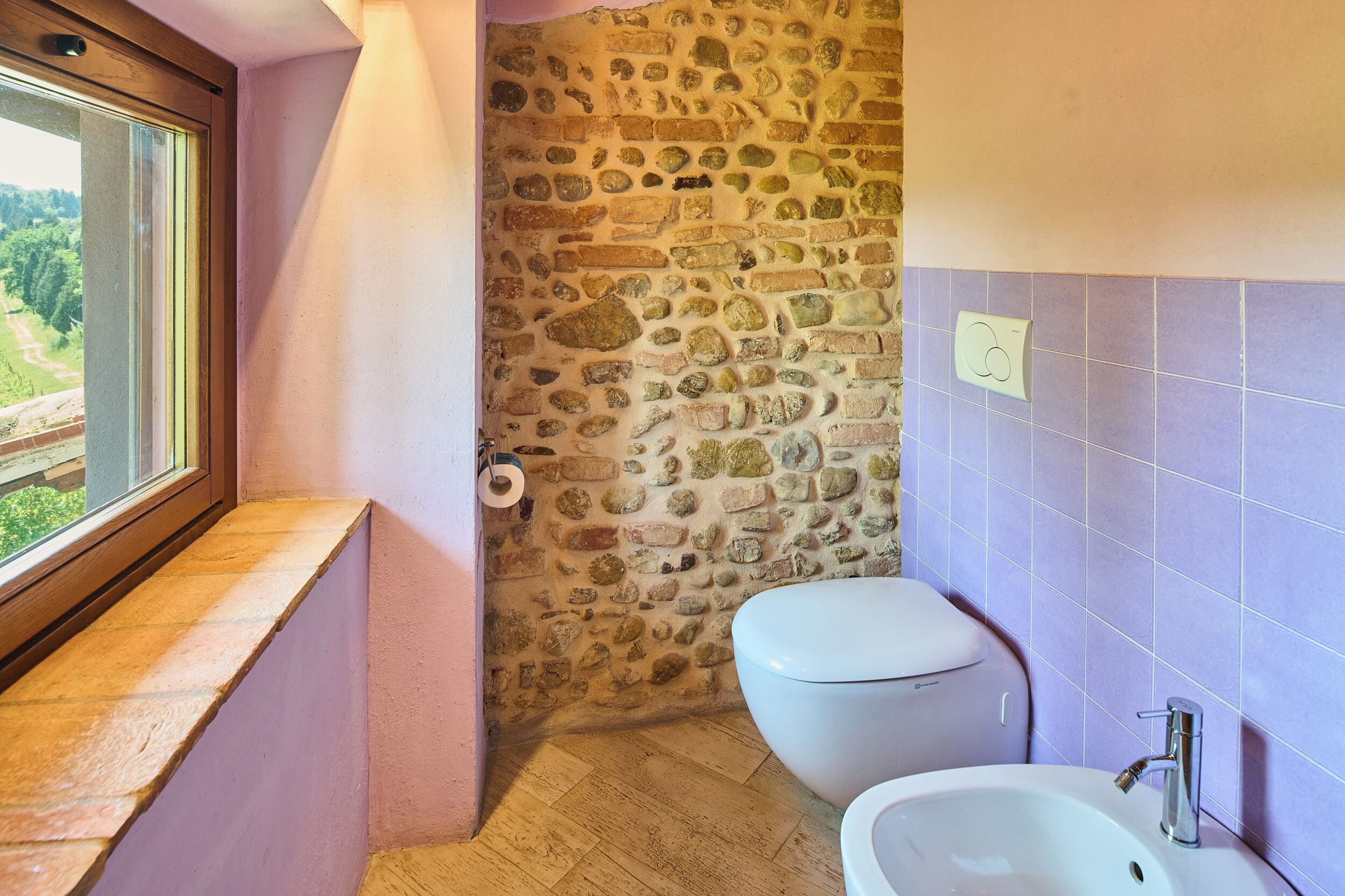 A bathroom with a stone wall, a toilet, a sink, and a window with a view of greenery.