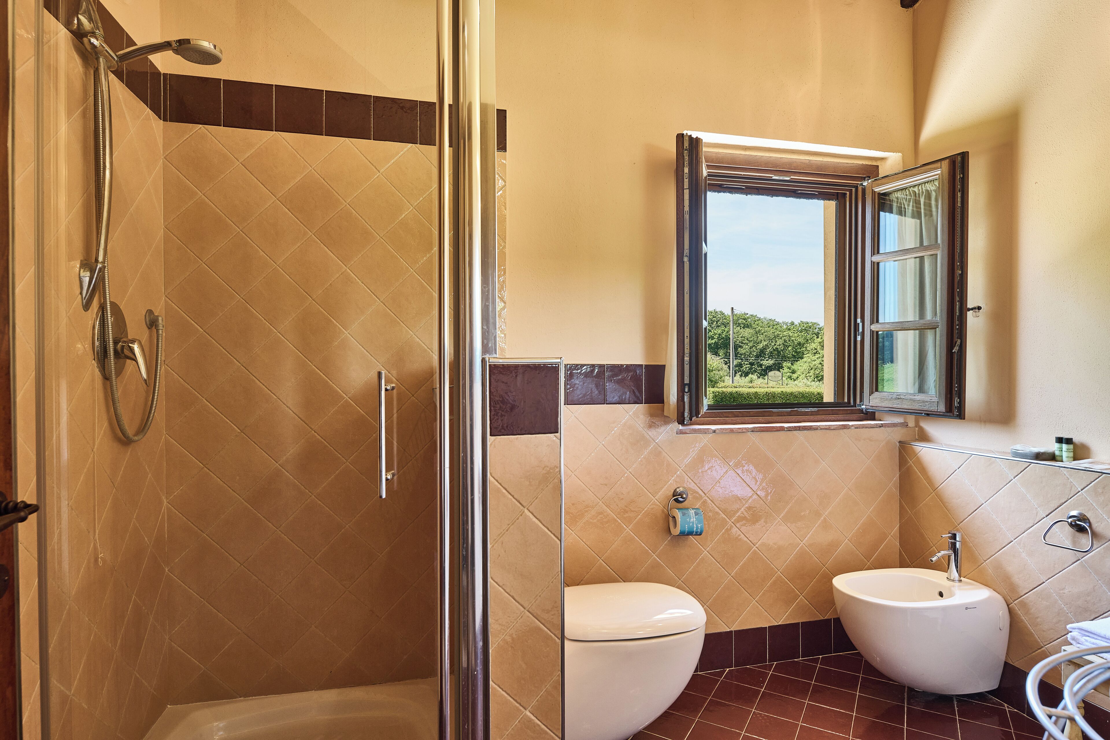 Une salle de bain équipée d’une douche, de toilettes, d’un bidet et d’une fenêtre donnant sur un espace verdoyant.