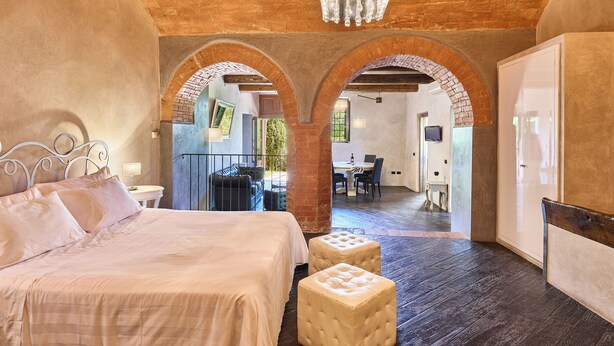 Een slaapkamer met een groot bed, twee voetenbankjes, een kroonluchter en een uitzicht door een boogvormige deuropening op een eetgedeelte.