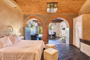 A bedroom with a large bed, two ottomans, a chandelier, and a view through an arched doorway to a dining area. - Borgo San Giusto (Empoli)