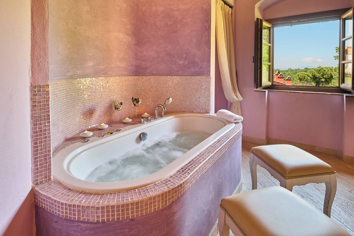 Une salle de bain avec une grande baignoire, une fenêtre donnant sur la verdure et un petit tabouret.