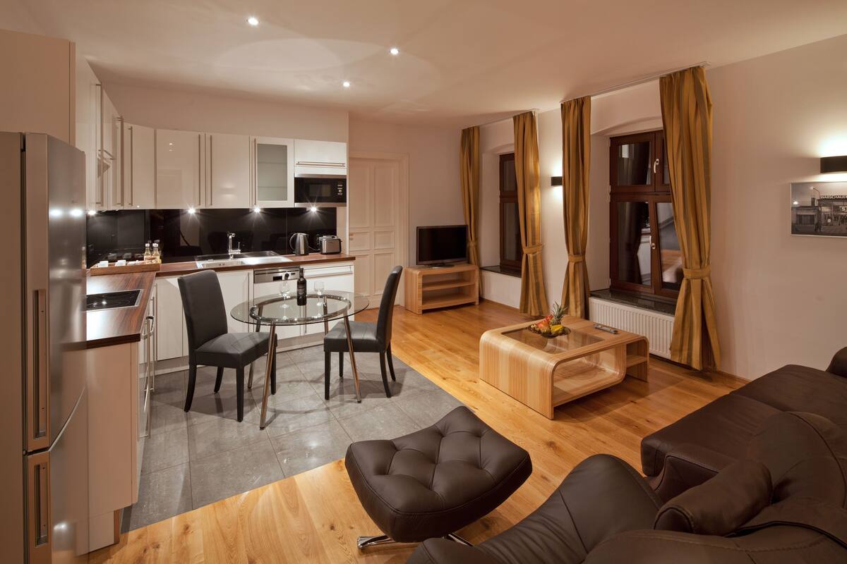 Una sala de estar moderna con un sofá, una mesa de centro y un comedor con una mesa de vidrio y sillas. Hay una cocina con refrigerador, microondas y estufa.