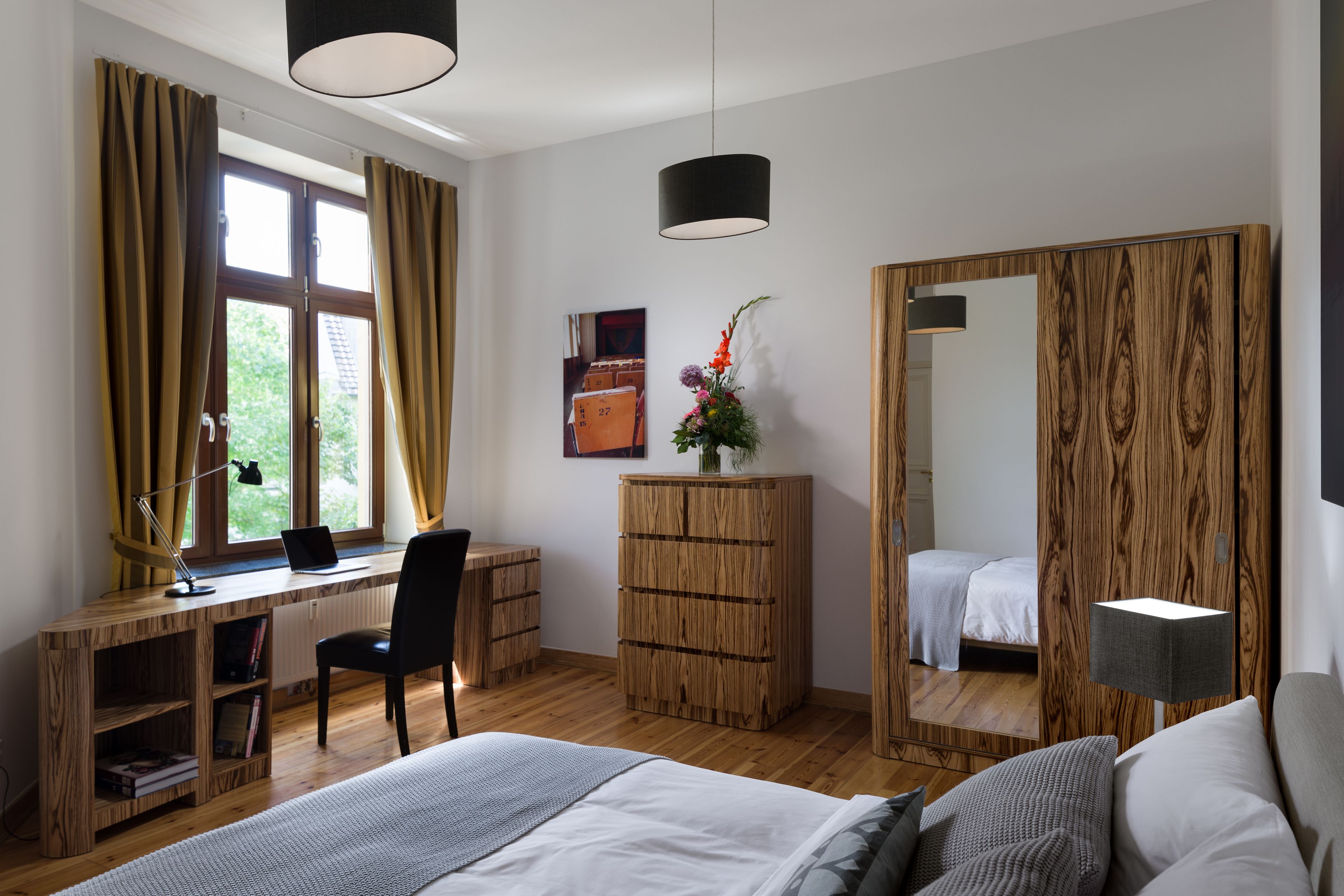 A bedroom with a wooden desk, a chair, a bed, a mirror, and a window with curtains.