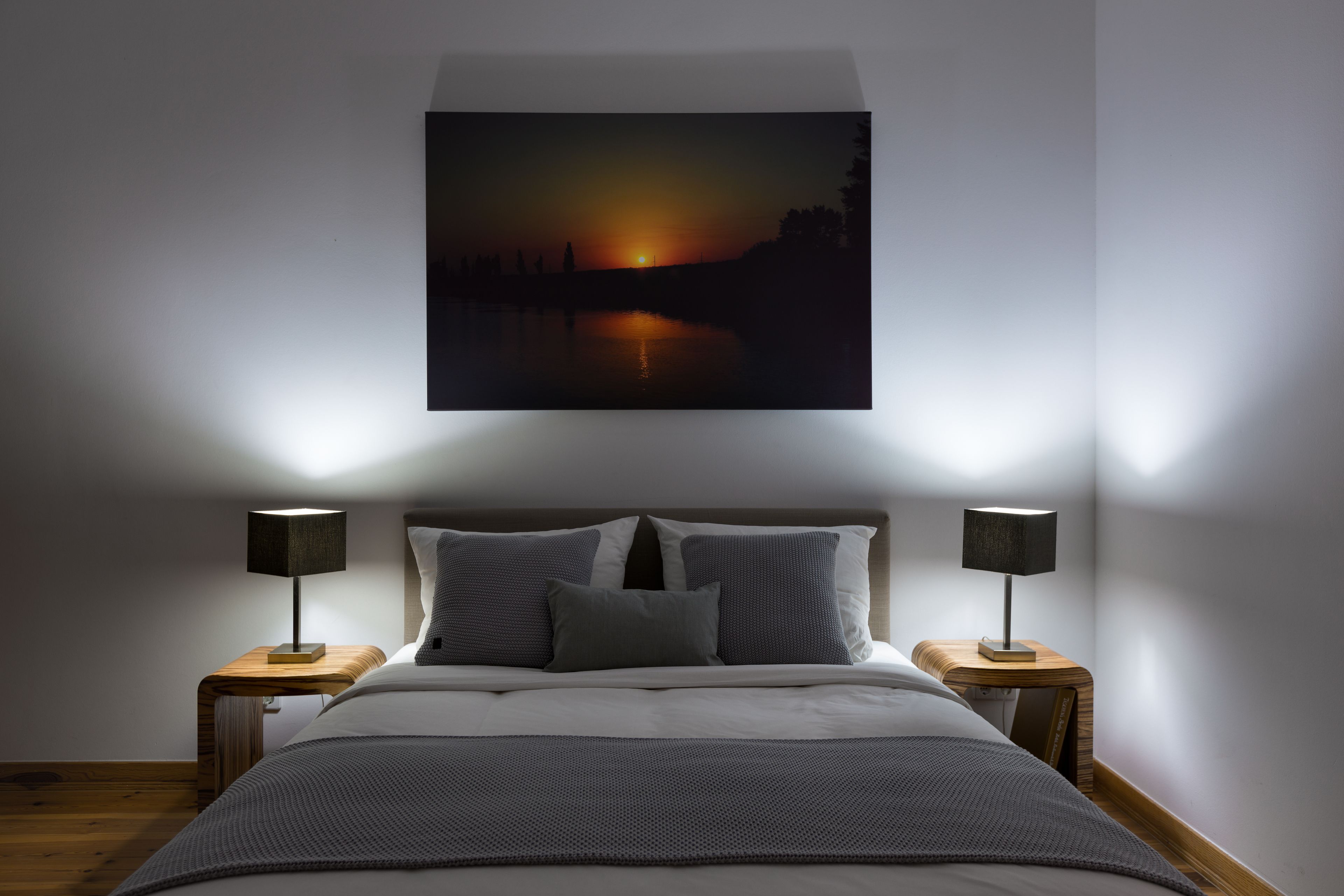 Une chambre à coucher comprenant un lit, deux tables de chevet et un tableau représentant un coucher de soleil sur une étendue d’eau, accroché au mur.