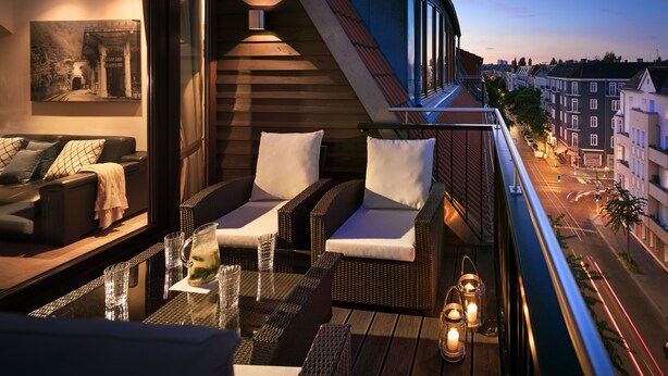 Een balkon met rieten meubilair, een glazen tafel en uitzicht op de stad in de schemering.