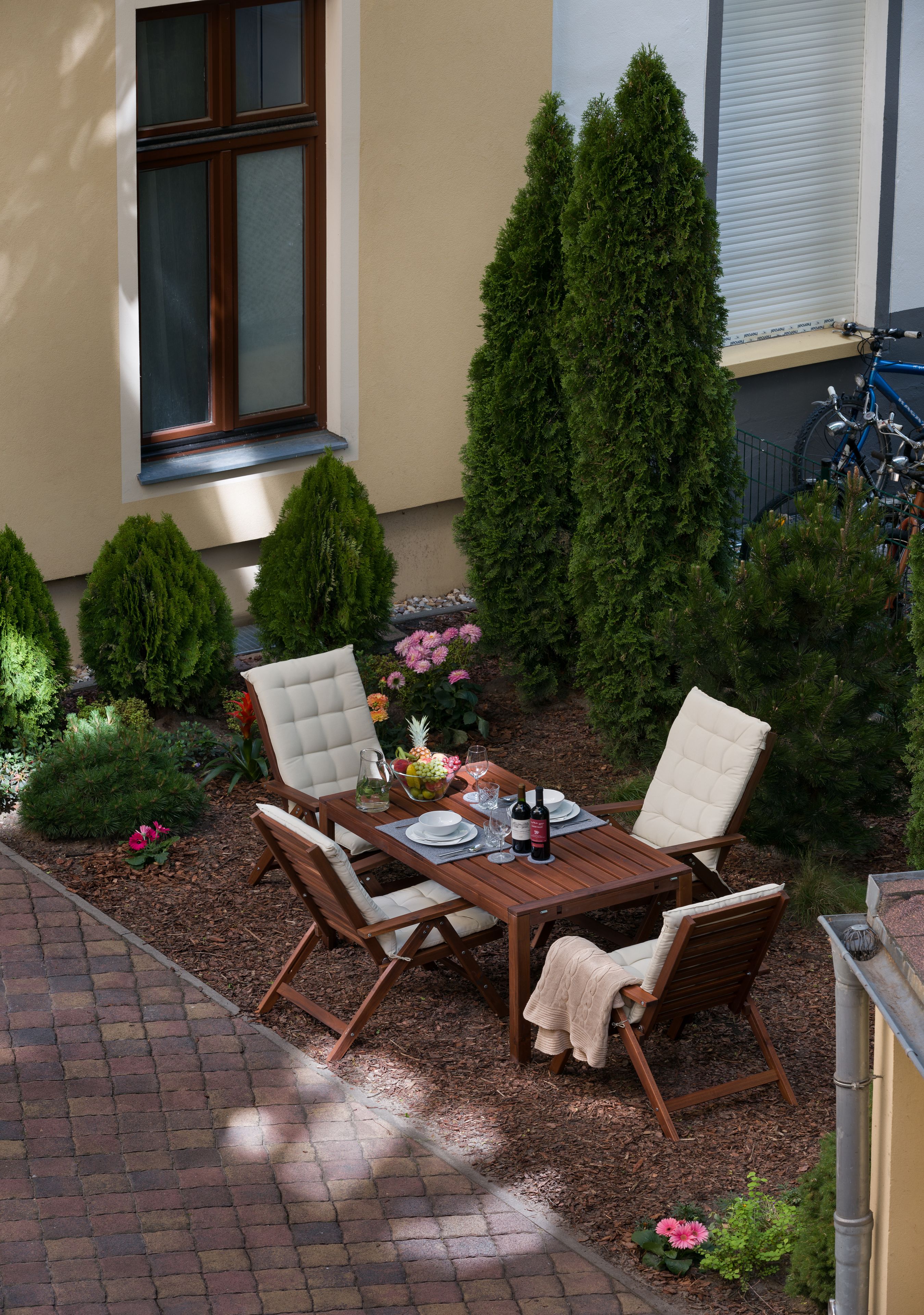 Eine Terrasse mit einem Holztisch und vier Stühlen, umgeben von Grünpflanzen und einem Fenster.