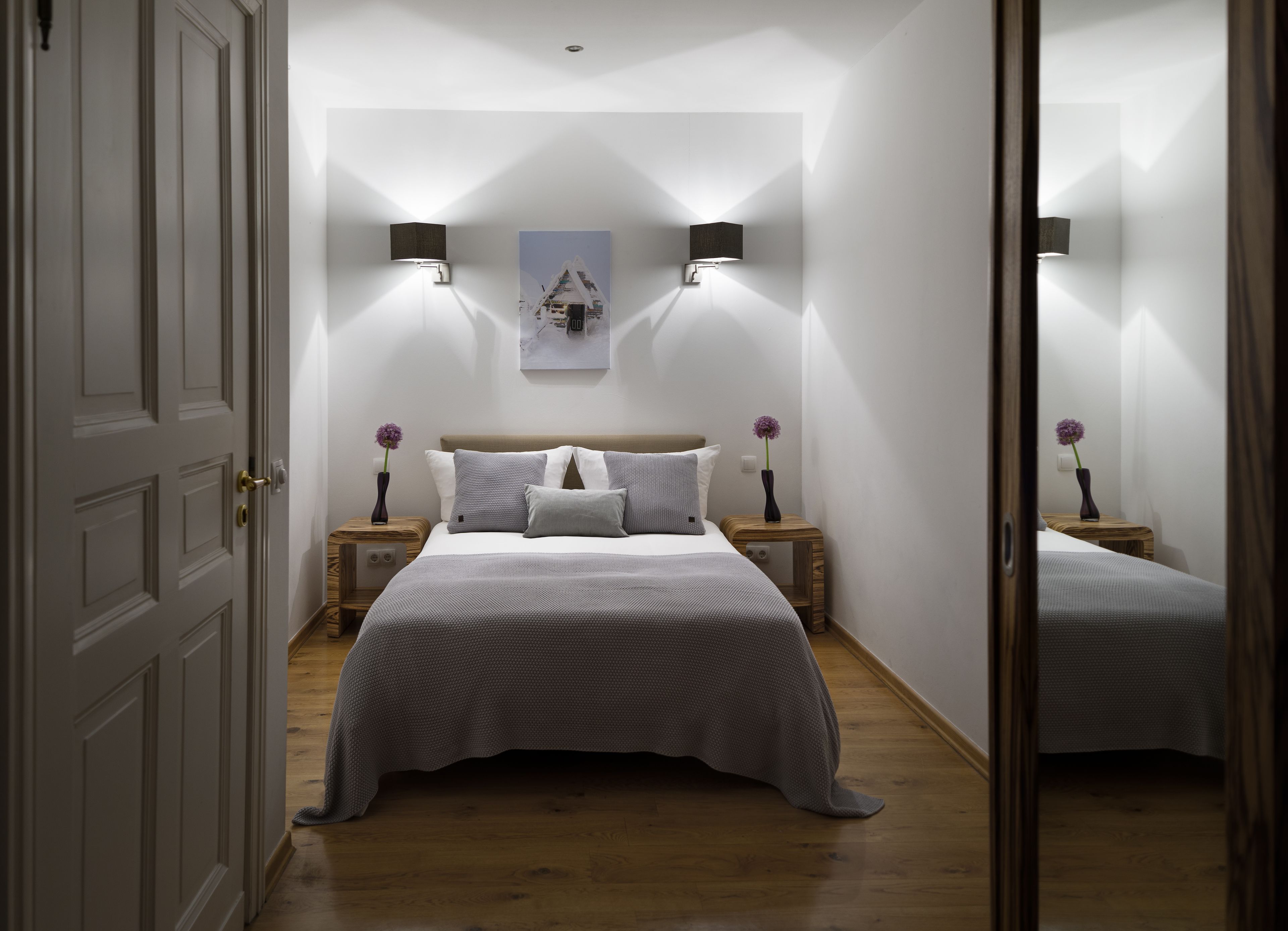 A bedroom with a bed, two bedside tables, a nightstand, a wall-mounted lamp, and a framed picture.