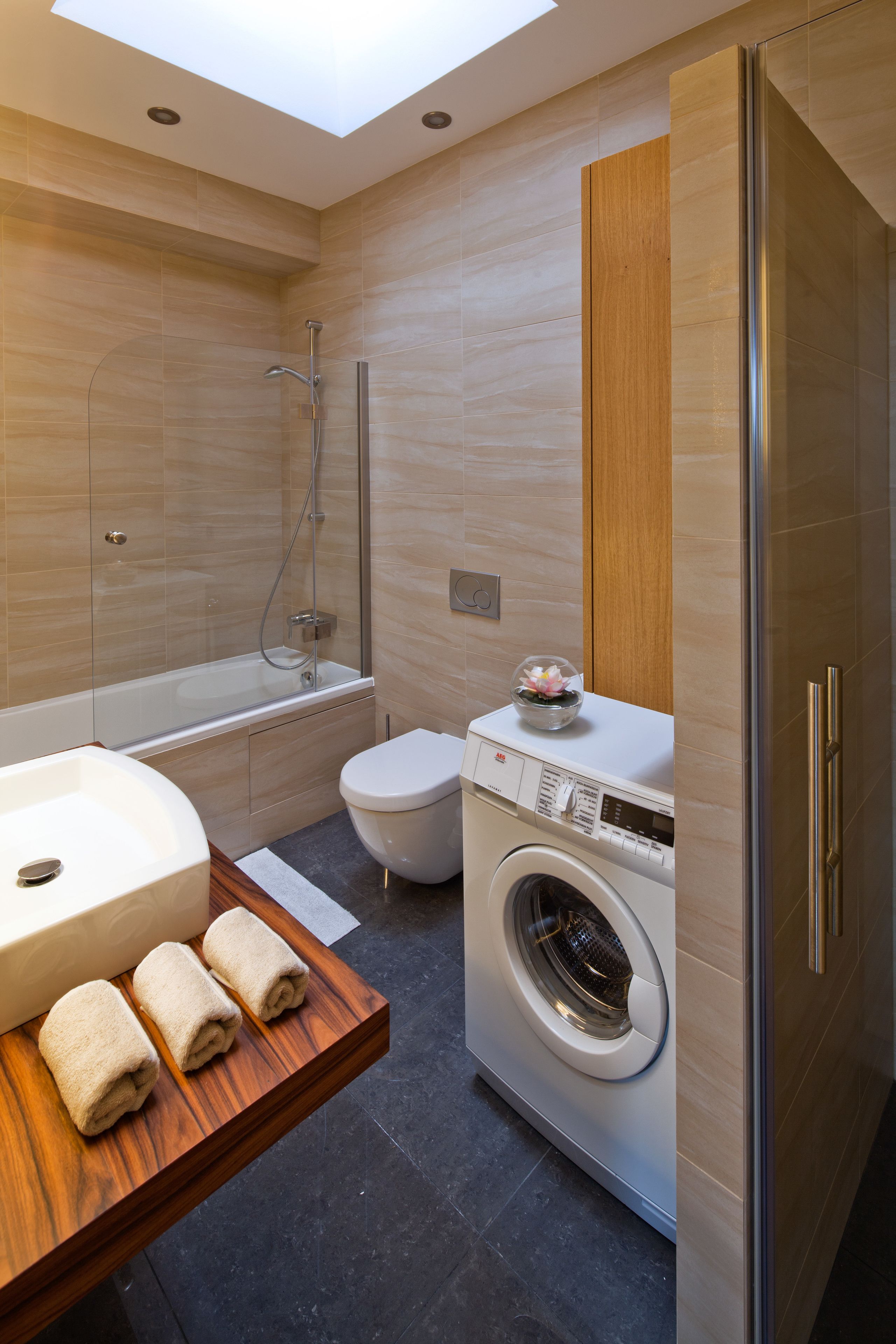 Een moderne badkamer met dakraam, ligbad, toilet, wasmachine en een houten wastafelmeubel met handdoeken.
