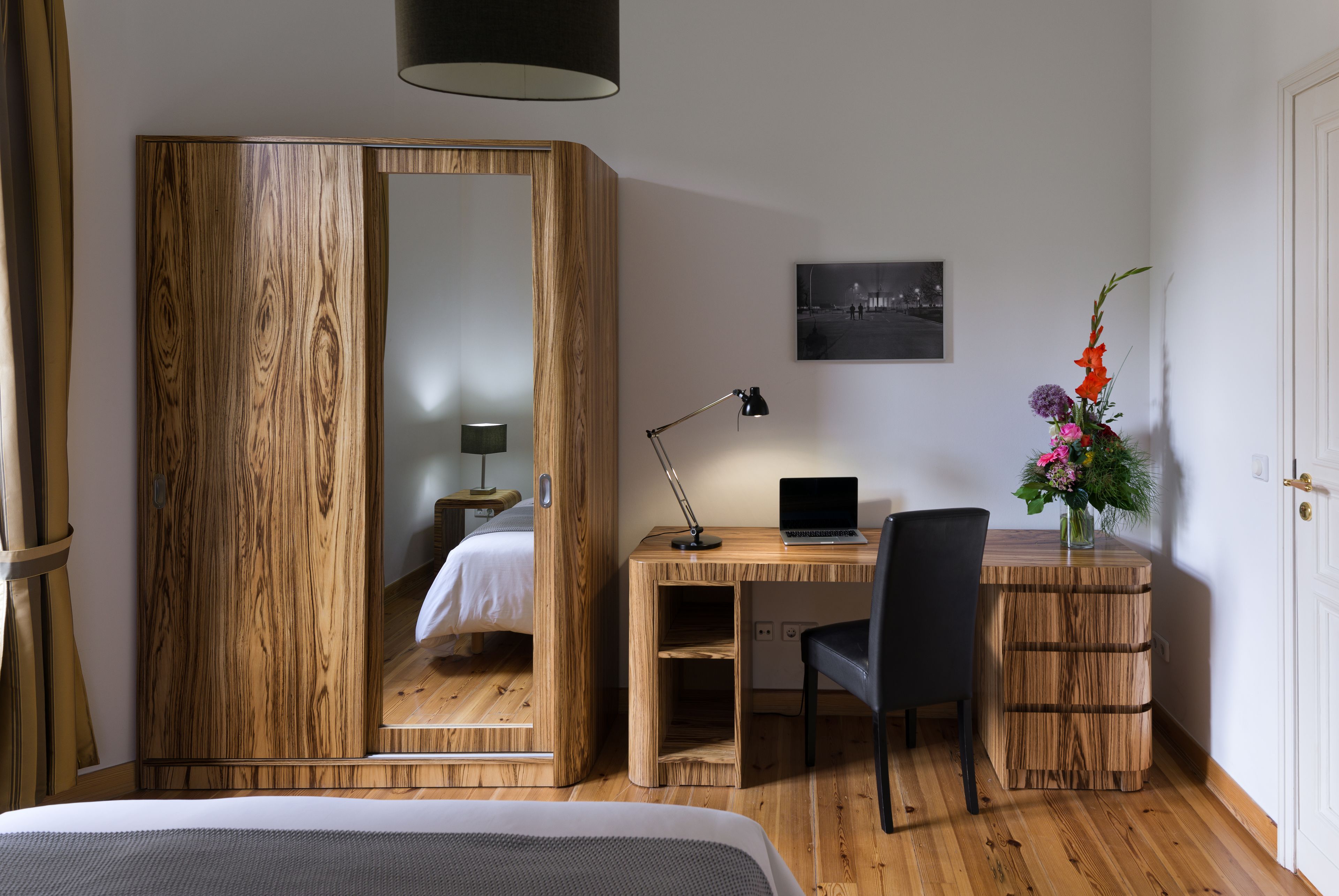 Un dormitorio con un armario de madera, un escritorio con una computadora portátil, una silla, una mesita de noche con una lámpara y un cuadro en la pared.