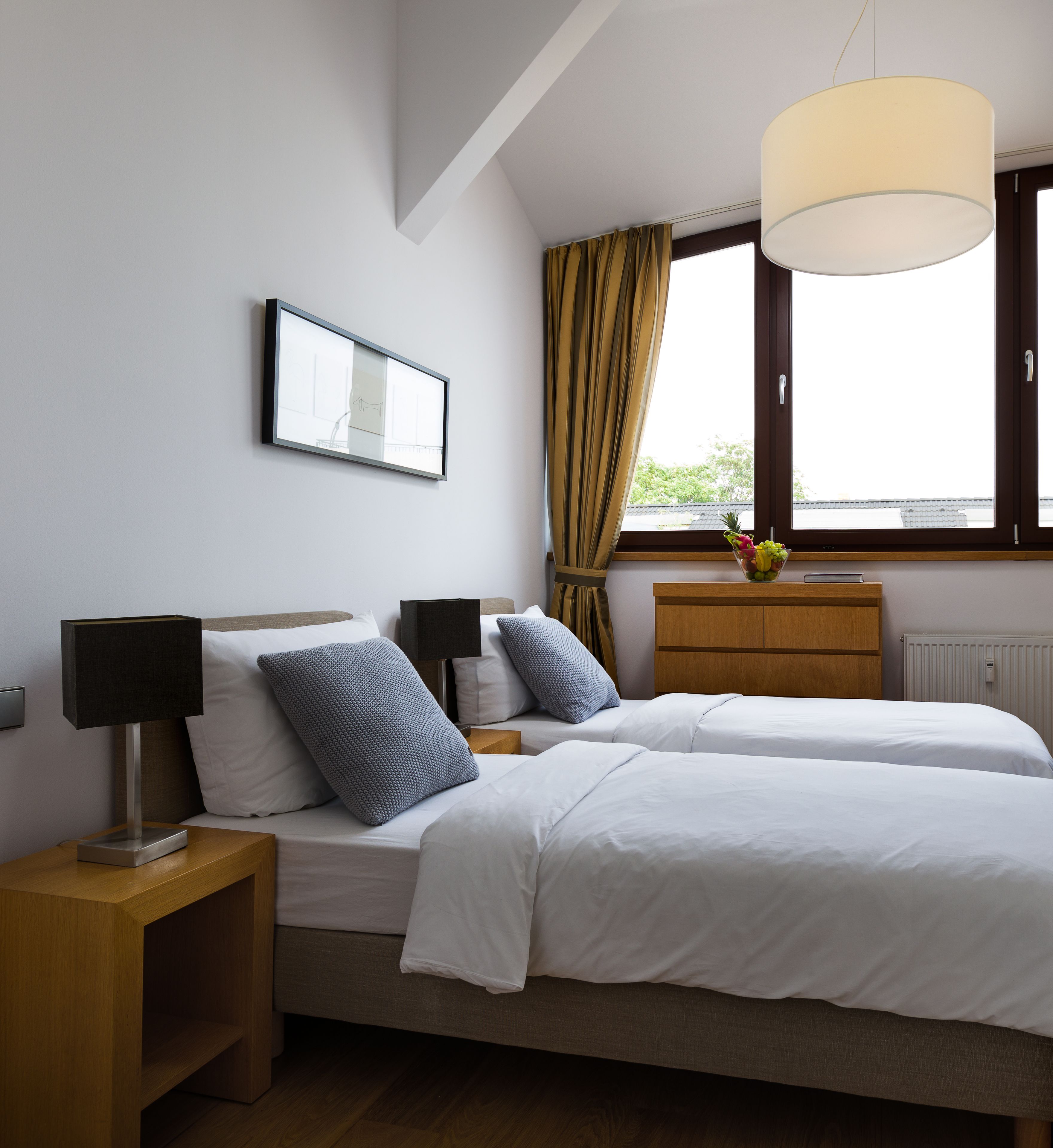 Um quarto com duas camas, uma mesinha de cabeceira com abajur, cômoda, janela com cortinas e um quadro na parede.