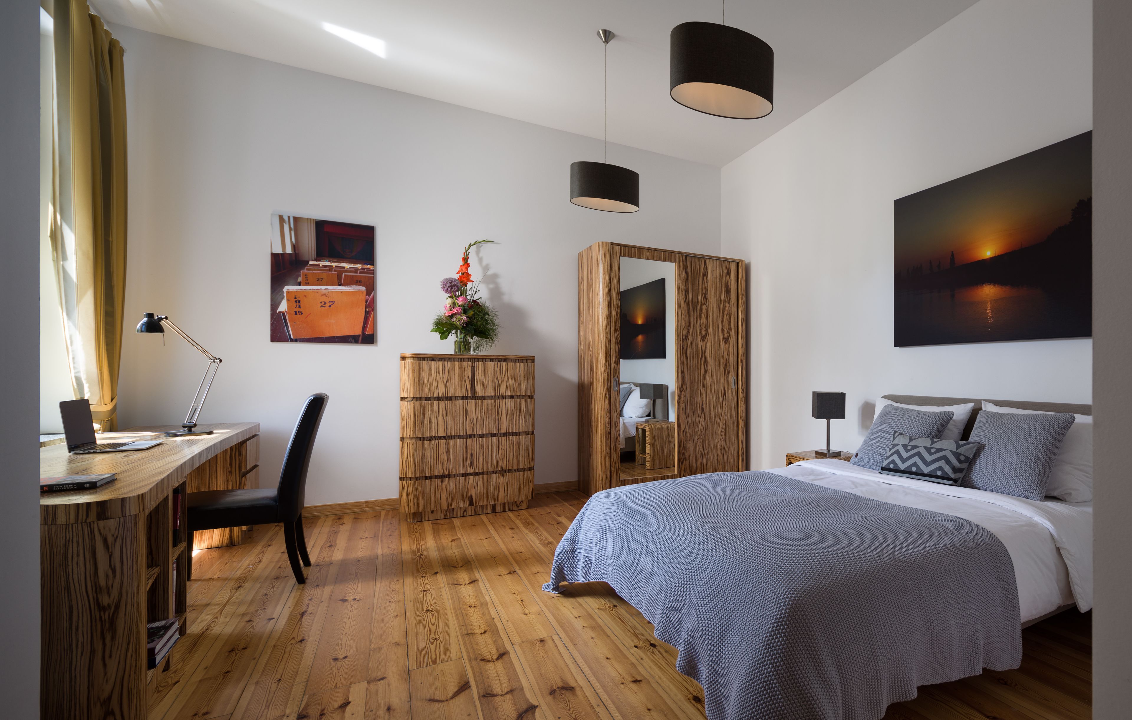 A bedroom with a wooden floor, a bed with a grey blanket, a desk with a laptop, a chair, a wooden wardrobe, and a painting on the wall.