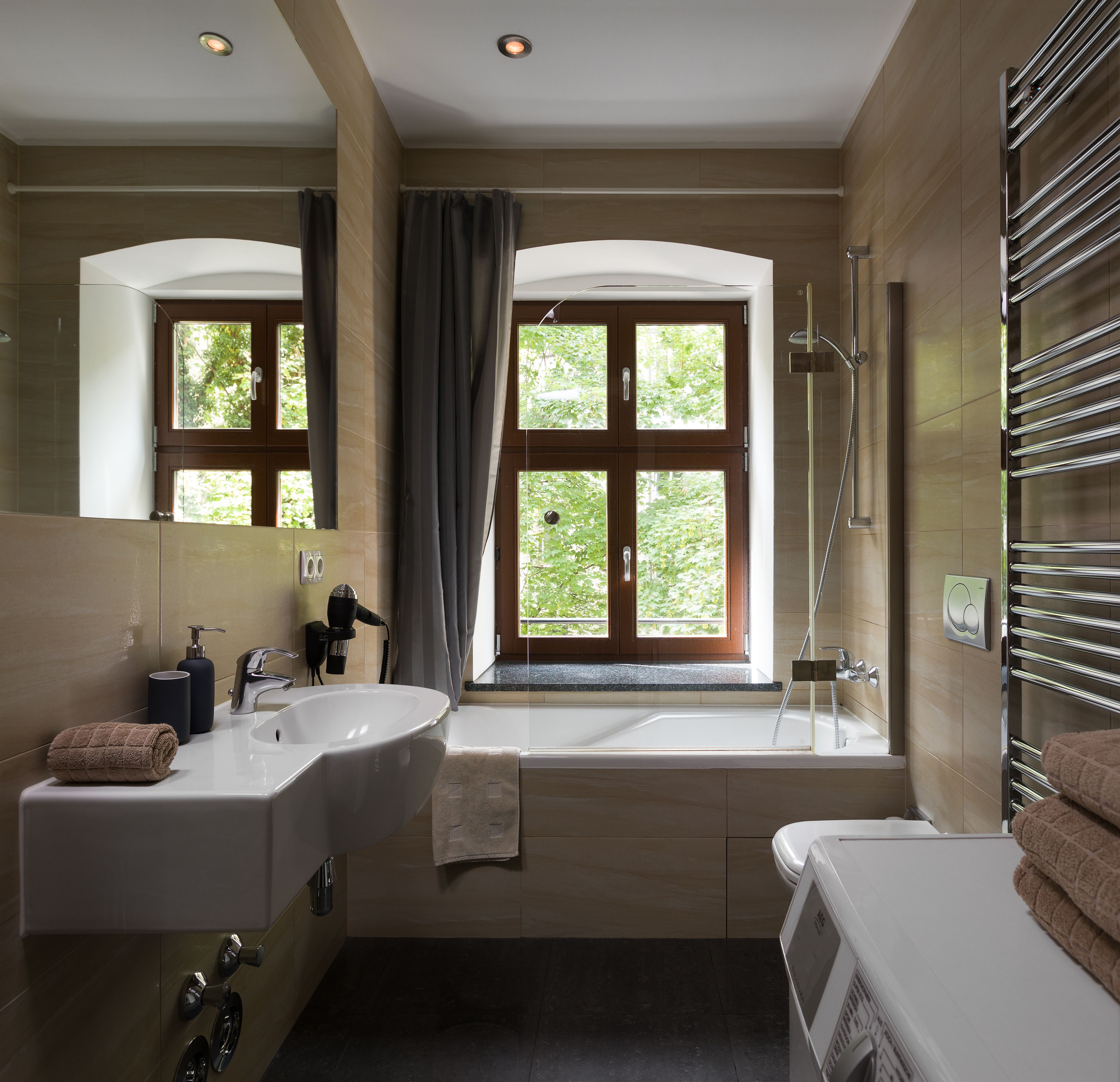 Un baño moderno con un lavamanos grande, una bañera y una ducha. Hay ventanas con cortinas, un perchero para toallas y un secador de pelo sobre el lavamanos.