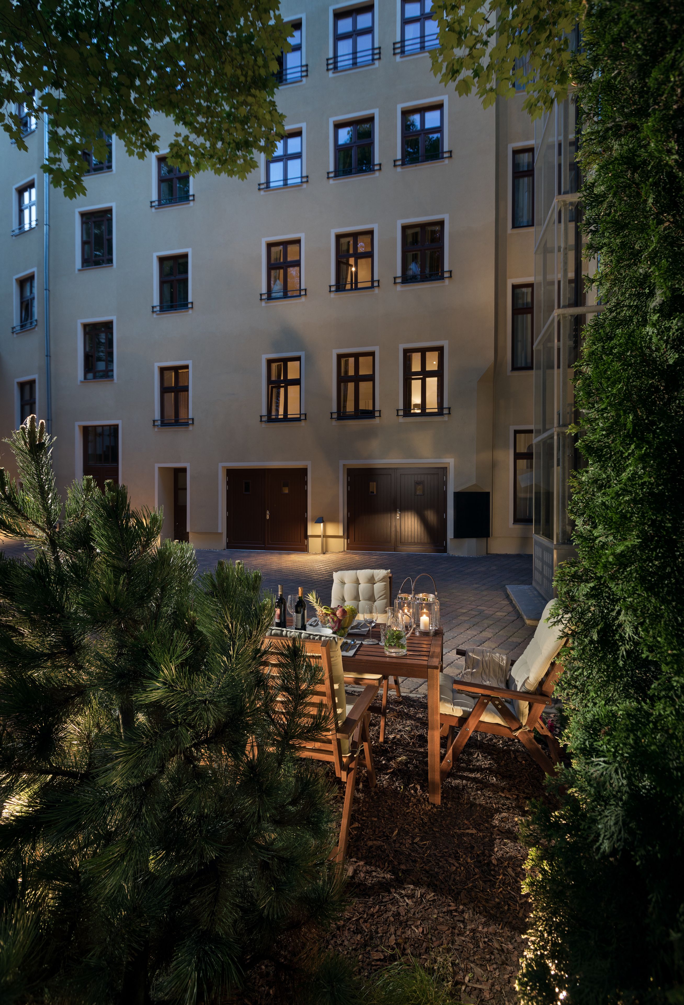 A courtyard with a wooden table set for dining, surrounded by pine trees and a building with multiple windows and doors.