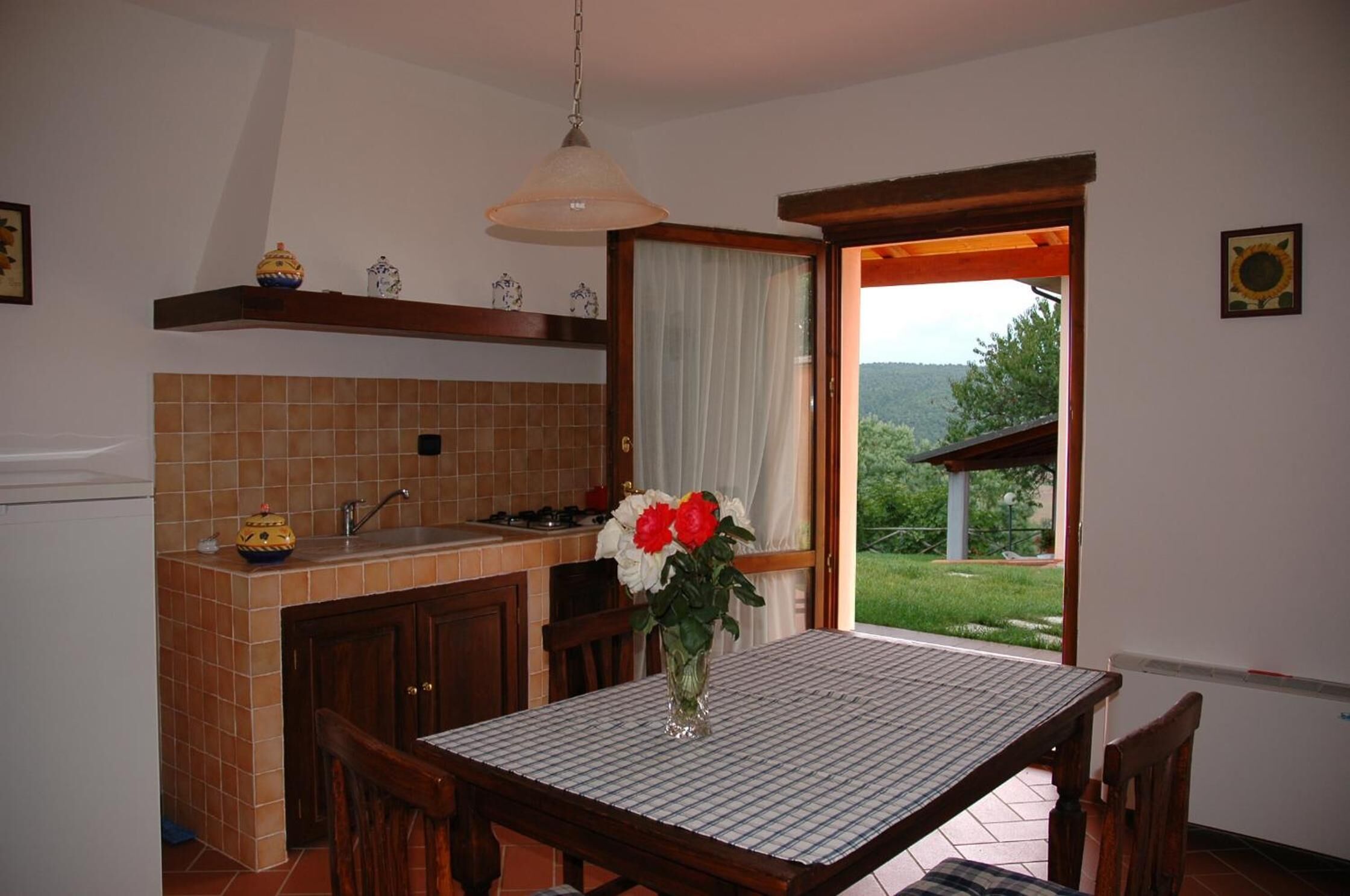 A kitchen with a tiled backsplash, a dining table with chairs, a vase with flowers, and a view of greenery through an open door.