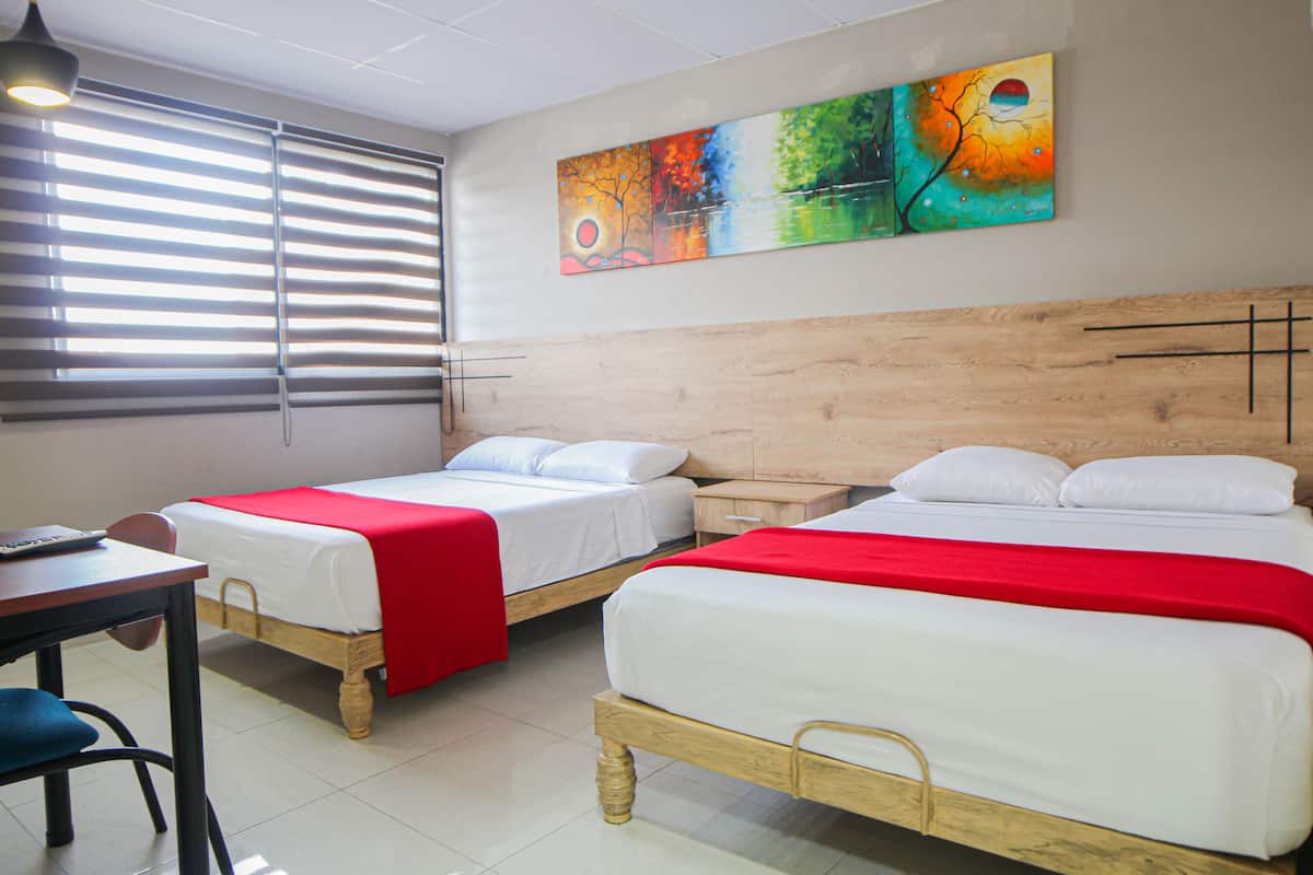 A hotel room with two single beds, a wooden headboard, a red and white bedspread, a wooden nightstand, a desk with a chair, and a painting on the wall.