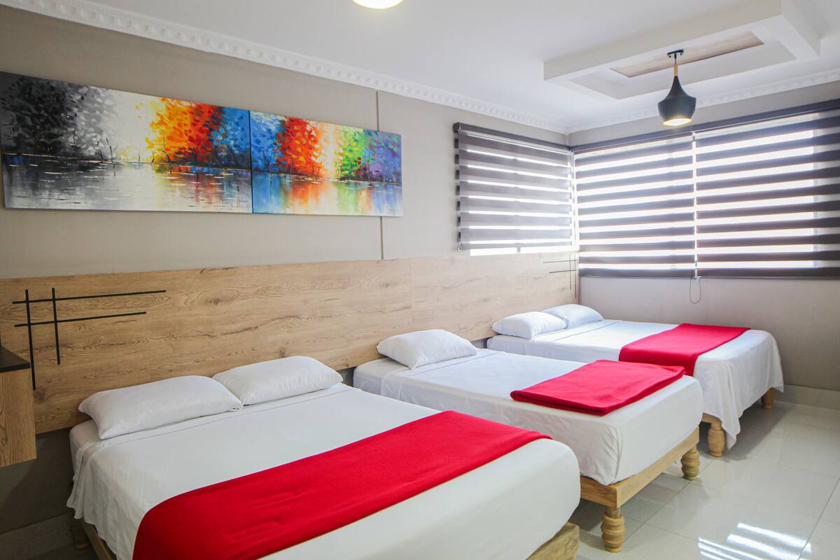 A hotel room with three beds, a wooden headboard, a colorful painting, and a window with blinds.
